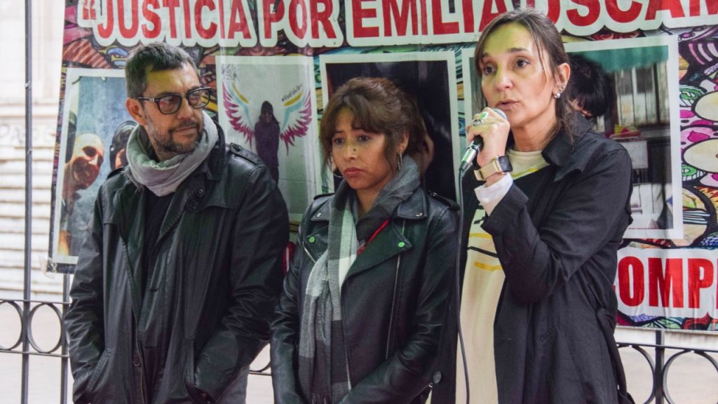 tres personas hablando ante un micrófono con un banner detrás que dice Justicia por Emilia